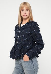 Jūras zila teksturēta jaka ar fringing detaļām, sudraba pogām un īsu dizainu, apvienota ar gaismas zila denim biksēm.