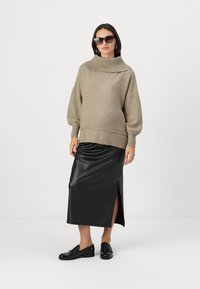 Pull en tricot beige surdimensionné avec un col montant et des poignets côtelés, associé à une jupe midi en cuir noir avec une fente latérale et des mocassins noirs.