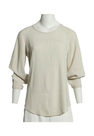 Beige pitkähihaisen puseron pyöreä pääntie, löysä istuvuus ja napilliset kalvosimet esiteltynä valkoisella päättömälla mallilla.