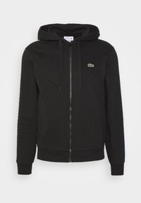 Svart zip-up hoodie i mjukt tyg med en figurnära design, med en känguroficka och en liten grön logotyp på bröstet.