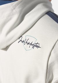 White cotton hoodie with a blue accent stripe and embroidered logo in dark blue and light blue, featuring a soft, textured fabric.