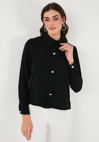 Black long-sleeve blouse with a pointed collar, front buttons featuring floral accents, and buttoned cuffs. Smooth texture, relaxed fit.