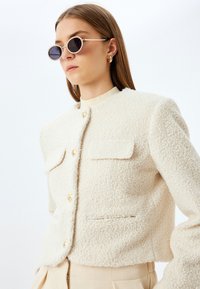 Veste courte de couleur crème avec une finition texturée et douce, ornée de boutons dorés et de deux poches avant. Lunettes de soleil rondes avec des verres sombres.