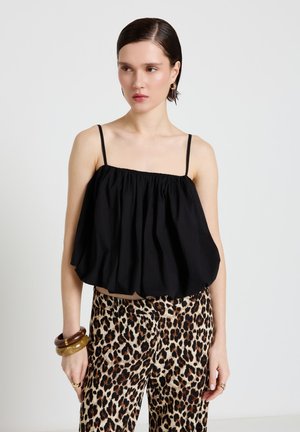 Donna con capelli lisci tirati indietro che indossa una canotta nera larga con spalline sottili, pantaloni con stampa leopardata e braccialetti spessi al polso sinistro.