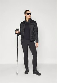 Γυναίκα με μαύρο μπουφάν, παντελόνι και παπούτσια, κρατώντας μπαστούνια πεζοπορίας, φορώντας μεγάλα μαύρα γυαλιά ηλίου, όρθια μπροστά σε απλή φόντο.