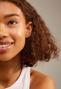 Pendientes de plata con un diseño texturizado que presenta formas abstractas. El modelo tiene el cabello rizado y castaño y lleva una blusa de cuello blanco.