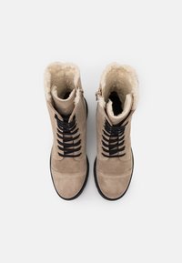 Béžové semišové kotníkové boty s černými šňůrkami, kožíškovou podšívkou a gumovou podrážkou, vybavené bočním zipem s malým zdobením na vrchu.