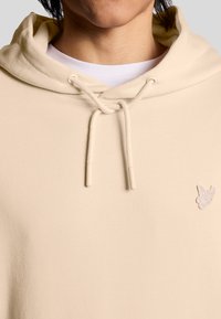 Beige Hoodie mit einer weichen Textur, ausgestattet mit einer Kordelzugkapuze und einem kleinen bestickten Logo auf der Brust. Weißes Shirt darunter sichtbar.