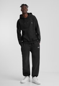 Felpa nera con cappuccio e lacci, dotata di tasca frontale, abbinata a pantaloni jogger neri con polsini elastici. Sneakers bianche completano l'outfit.