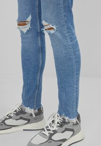 Jeans decoloridos en azul claro con dobladillos deshilachados y roturas en las rodillas, combinados con zapatillas grises y blancas que presentan un diseño de malla y ante.