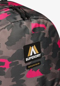 Mochila de camuflaje negra y rosa con una superficie texturizada, que presenta el logo de Superdry y una etiqueta que dice "MOUNTAIN", cremalleras dobles y correas acolchadas.