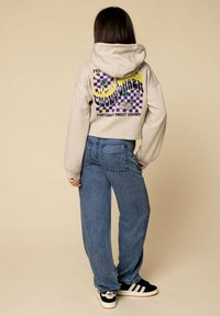 Beige cropped hoodie met kleurrijke grafische print op de achterkant, gecombineerd met losse blauwe denim jeans en zwarte sneakers met witte strepen.