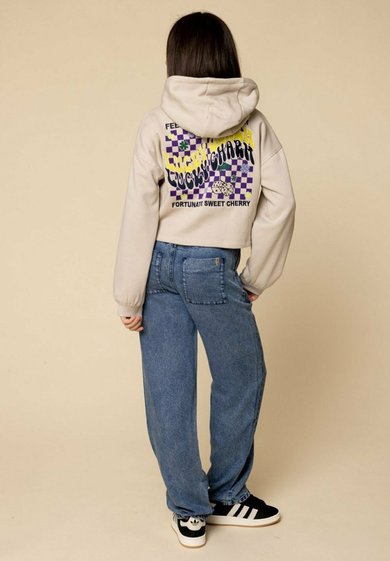 Beige cropped hoodie met kleurrijke grafische print op de achterkant, gecombineerd met losse blauwe denim jeans en zwarte sneakers met witte strepen.
