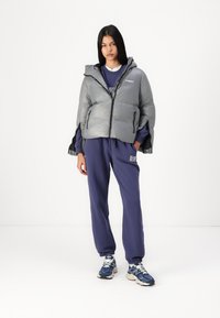 En puffjakke i metallisk grå med glidelås foran, hette og sidelommer, sammen med marineblå sweatpants og flerfargede joggesko.