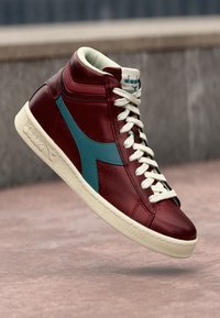 High-top sneaker in burgundy leather with teal accents, white laces, and a textured cream sole; streamlined design with stitched details.