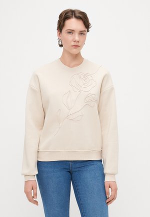 Sweatshirt bege com gola redonda, apresentando um desenho de rosa bordado na frente. Tecido macio e punhos canelados, combinado com jeans azuis.