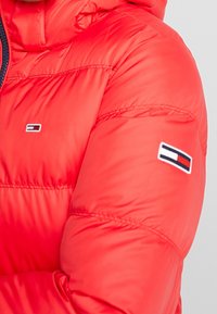 Rød pufferjakke med lynlås foran, hætte og et logo-patch på ærmet. Materialet er glat med en quiltet tekstur.