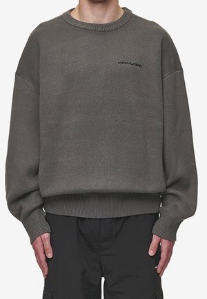 Uomo che indossa un maglione grigio a tessuto spesso e vestibilità ampia, con polsini e orlo a coste, abbinato a pantaloni neri, in piedi con le mani rilassate ai lati.