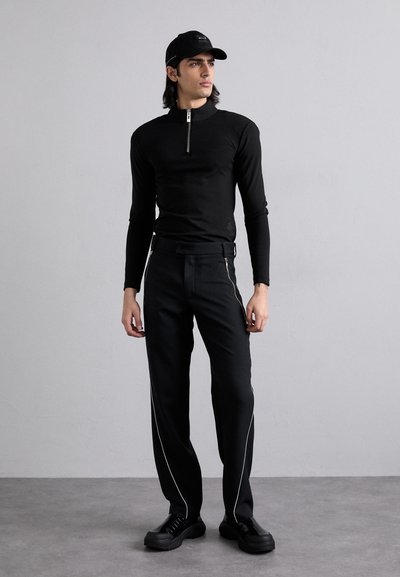 Top noir à manches longues avec un col zippé, associé à un pantalon noir avec des zips sur les côtés et des passepoils blancs, et des chaussures noires à lacets.