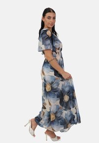Blomstret chiffon maxikjole med store blå og beige blomsterprints, korte flæser ved ærmerne og en tætsiddende talje, båret med hvide hæle.