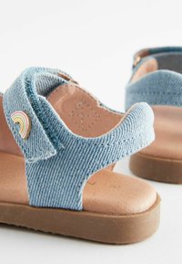 Denimsandaler med överdel i ljusblått tyg, brun texturerad sula och en regnbågsaccent på remmen. Justerbar rem för passform.