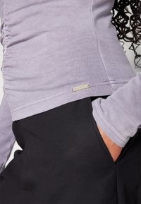 Lavendelfarbenes Langarmoberteil mit gerafftem Seitendetail, kombiniert mit schwarzen Hosen, die eine Seitenasche haben. Silberner Markenanhänger am Saum.