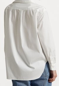 Takaisin näkyy henkilö, joka on pukeutunut löysään valkoiseen pitkähihaiseen paitaan, jossa on laskostustiedot, yhdistettynä sinisiin farkkuihin yksinkertaista taustaa vasten.