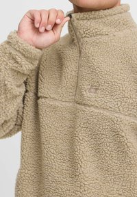 Beiger Fleece-Pullover mit einer strukturierten, flauschigen Oberfläche. Mit einem halb gezippten Hals und dezentem Logo-Detail auf der Brust.