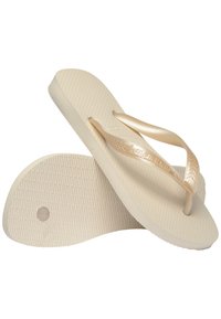 Beige papucsok texturált talppal és fényes világos bézs pántokkal, enyhe ívvel és középső lábujj párnával a láb támogatásáért.