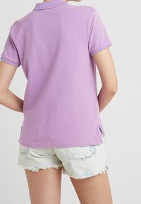 Fialová polo košile s texturovaným povrchem, krátkými rukávy a bočními rozparky, spárovaná s roztrhanými světlými denimovými šortkami.