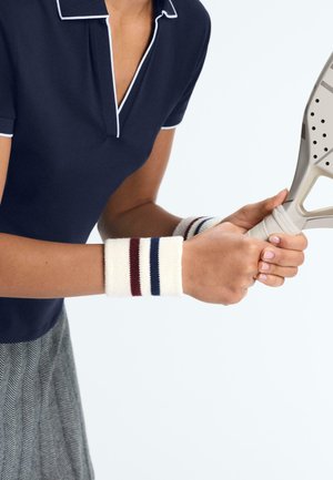 Personne portant un polo bleu marine et des bracelets rayés tenant une raquette de paddle beige sur un fond clair uni.