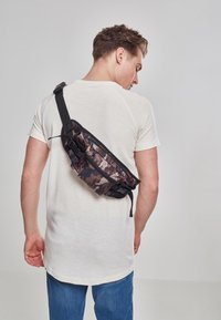 Borsa a tracolla mimetica realizzata in tessuto resistente, con scomparto con zip e spallaccio regolabile, indossata sulla schiena di una persona.