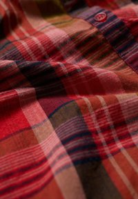 Tkanina w kratę z czerwonymi, różowymi, zielonymi i niebieskimi liniami i teksturowanym wykończeniem, z widocznym guzikiem blisko kołnierza.