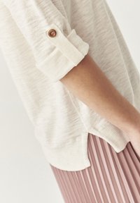 Helles beiges Sweatshirt mit aufgekrempelten Ärmeln und einem Holzkneippdetail, kombiniert mit einem plissierten rosa Rock; weiche Textur und lässige Passform.