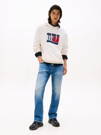 Beige Kapuzenpullover mit Logo-Druck, kombiniert mit hellblauen Jeans. Die Schuhe sind schwarze Sneaker mit strukturierten Details.