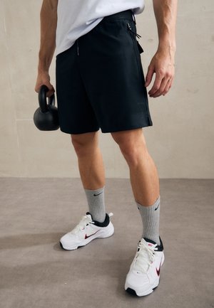 Mann in schwarzen Shorts, weißen Sneakern und grauen Nike-Socken hält eine schwarze Kettlebell auf einem grauen Boden vor einem hellbeigen Wandhintergrund.