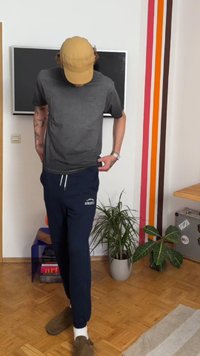 Gray short-sleeve t-shirt with a logo, navy sweatpants featuring white text "Athletic," and tan cap worn backward, standing on wood flooring.