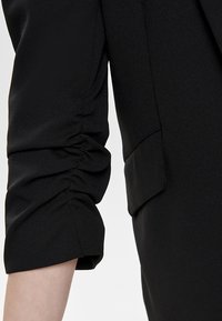 Blusa negra con mangas fruncidas, corte entallado, textura suave y detalle de bolsillo lateral. La tela parece ligera.