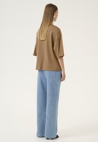 Hnědý bavlněný oversized top s detaily v oblasti ramene, spárovaný se světle modrými širokými džíny, s jemnými texturami a kapsami.