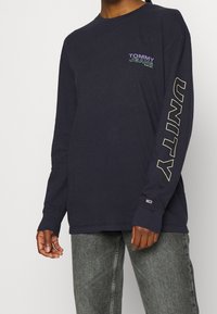 Marineblå långärmad skjorta i bomull med "TOMMY JEANS"-logotyp i lila och grön, samt "UNITY" tryckt i stora bokstäver längs ärmen.