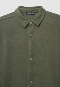 Cardigan verde oliva a maglia con collo a camicia, cinque bottoni sul davanti e un tessuto texturizzato. L'etichetta reca la scritta "CALLIOPE."
