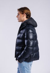 Sort pufferjakke lavet af blank læder, med hætte, lynlåsafslutning og quiltet design. Bæres med lyseblå jeans.