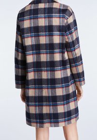 En person som bär en knälång ullrock med en beige, marinblå, blå och röd rutig mönster, stående mot en neutral vit bakgrund.