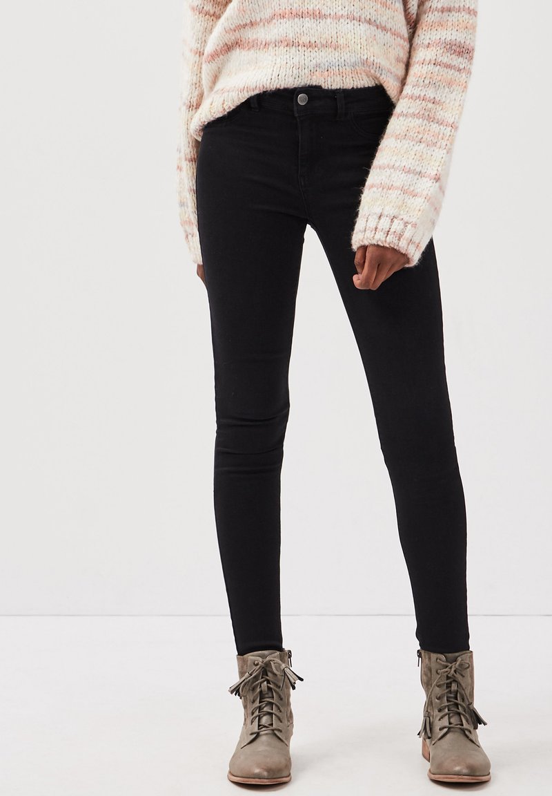 Jeans ajustados negros con una textura suave, cintura alta y diseño estándar de cinco bolsillos, combinados con botas de tobillo ligeras de color gris claro con cordones.