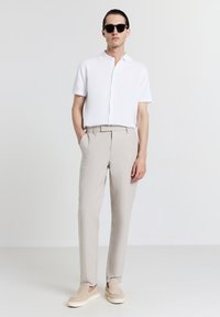 Weißes Kurzarm-Hemd mit Knöpfen, beigefarbene gestreifte Hose und Slipper. Eine minimalistische Sonnenbrille rundet das Outfit ab.