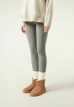 Person trägt graue gerippte Leggings, cremefarbenen übergroßen Pullover, weiße geraffte Socken und braune Wildleder-Stiefeletten vor hellem Hintergrund.