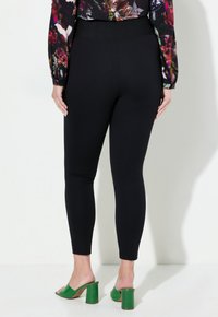 Mustat legginsit, joissa on korkea vyötärö ja virtaviivainen, tiukka muotoilu. Materiaali on sileä ja siinä on hienoinen kiilto. Jalkineena on vihreät korkokengät.