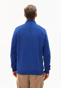 Blauwe sweatshirt met hoge kraag, zachte textuur, ribgebreide manchetten en zoom, gedragen over lichtbruine corduroy broek. Achteraanzicht toont een eenvoudig ontwerp.