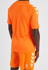 Camiseta atlética naranja y pantalones cortos hechos de material liviano. Tiene acentos en chevrón blanco en las mangas y los pantalones cortos, con detalle de logotipo.