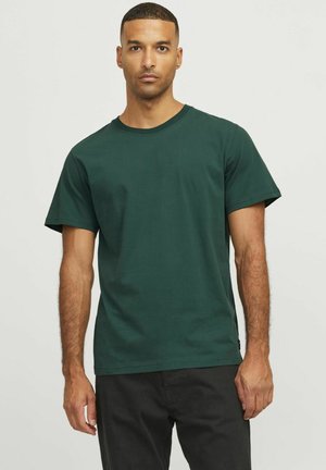 Mann mit kurzen Haaren, der ein einfaches dunkelgrünes T-Shirt mit kurzen Ärmeln und schwarze Hosen trägt, steht vor einem hellen Hintergrund.